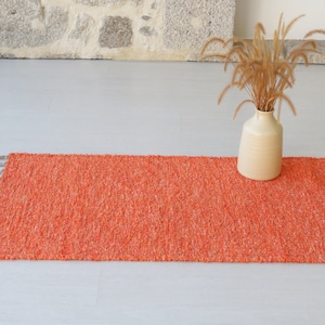 May include: A rectangular orange rug with a textured surface and fringed edges. The rug is approximately 91 cm long and 51 cm wide. A beige vase with dried plants sits on the right side of the rug.