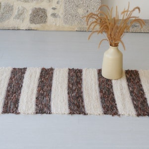 May include: A rectangular rug with cream and brown vertical stripes. The rug has a fringed edge. A beige vase with dried ornamental grass sits on the rug. The rug is on a light grey surface.