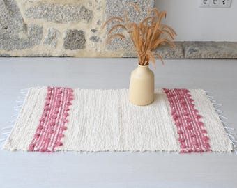 Può includere: Un tappeto rettangolare bianco sporco con dettagli rosa. Il tappeto presenta una trama strutturata e strisce verticali rosa in rilievo su ciascuna estremità. Un vaso beige con piante essiccate è posto sul tappeto.
