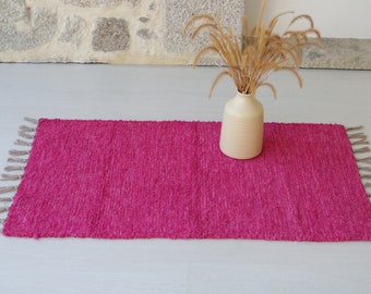 Op de afbeelding: Een roze geweven tapijt met franjes aan de uiteinden. Het tapijt ligt op een witte vloer en een vaas met gedroogd gras staat op de achtergrond.