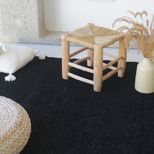 May include: A black woven rug with a natural woven stool and two white throw pillows. A vase with dried grass sits on the rug.