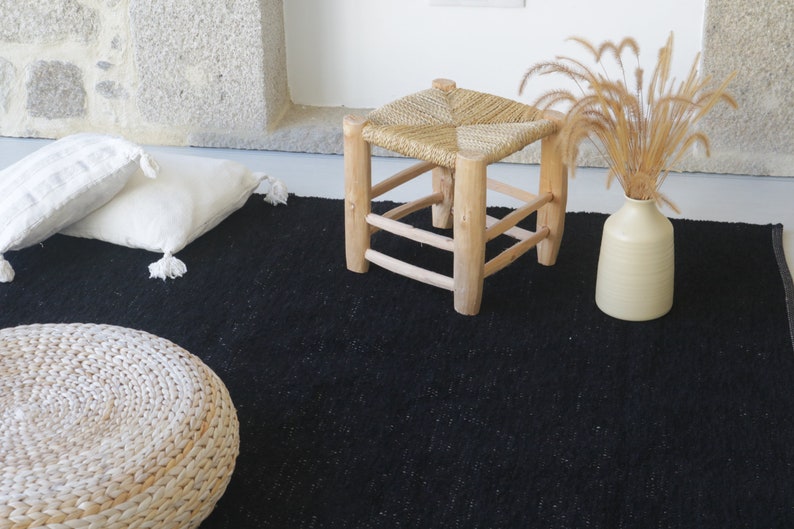 May include: A black woven rug with a natural woven stool and two white throw pillows. A vase with dried grass sits on the rug.
