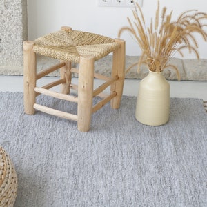 May include: A gray woven rug with fringe edges, a woven stool with a square top, and a woven round ottoman. The rug is on a light wood floor.