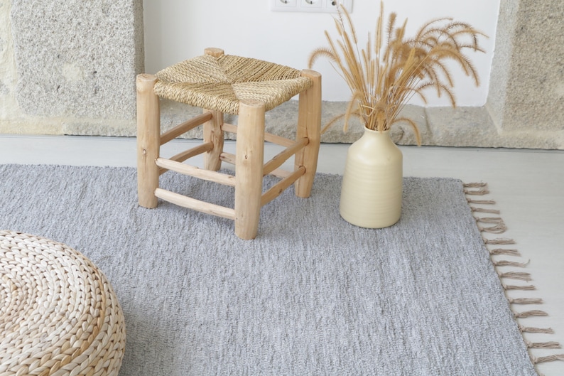 May include: A gray woven rug with fringe edges, a woven stool with a square top, and a woven round ottoman. The rug is on a light wood floor.