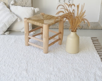 May include: A white fluffy rug with fringe edges, a woven stool with a wooden frame, and a vase with dried grass.