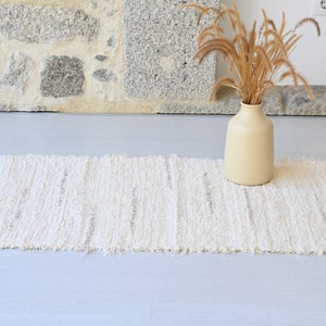 May include: A white woven rug with a fringed edge, placed on a light gray floor. A beige ceramic vase with dried grass is in the background.