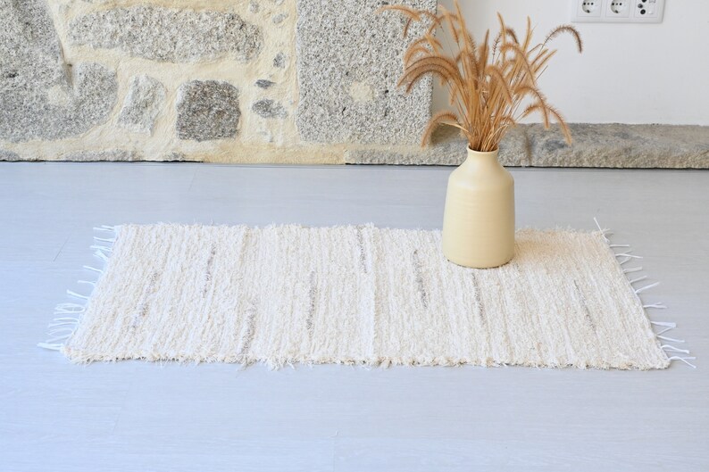 May include: A white woven rug with a fringed edge, placed on a light gray floor. A beige ceramic vase with dried grass is in the background.