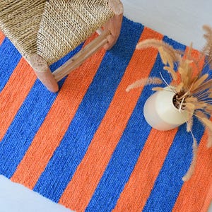 May include: A rectangular woven rug featuring bold, alternating vertical stripes in vibrant orange and royal blue. The rug has a textured, knotted surface and is displayed with a woven straw stool and a white vase containing dried grasses.