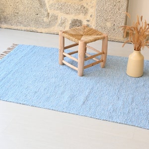 Pu&ograve; includere: Un tappeto blu chiaro con frange sui bordi. Il tappeto &egrave; su un pavimento bianco e uno sgabello di legno con una seduta intrecciata &egrave; al centro del tappeto.