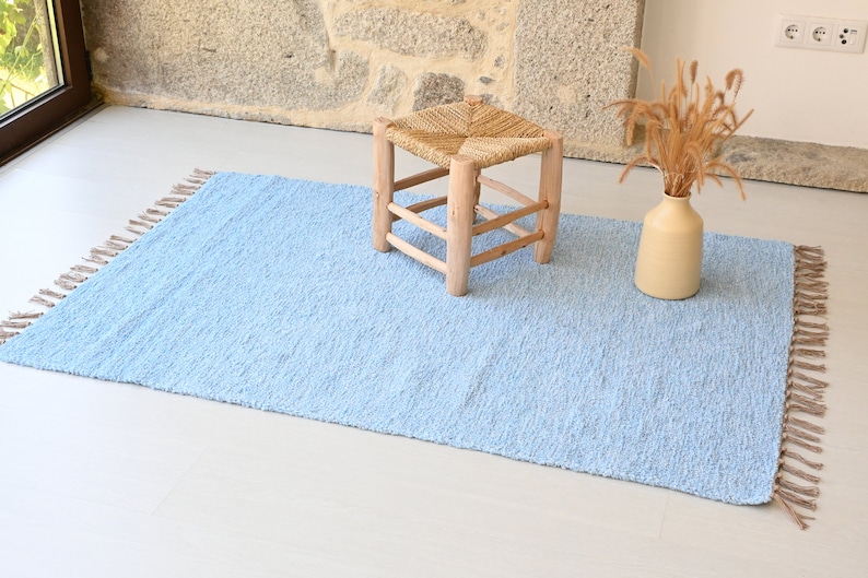 Pu&ograve; includere: Un tappeto blu chiaro con frange sui bordi. Il tappeto &egrave; su un pavimento bianco e uno sgabello di legno con una seduta intrecciata &egrave; al centro del tappeto.