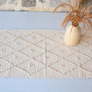 Op de afbeelding: Crèmekleurig rechthoekig tapijt met een ruitpatroon en getextureerde details. Een beige vaas met gedroogde planten staat op het tapijt. Het ontwerp van het tapijt heeft verhoogde, gestippelde accenten.
