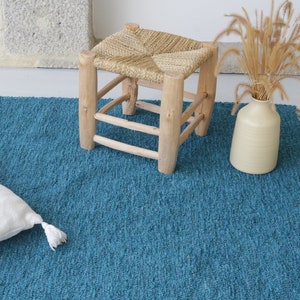 May include: A teal blue textured rug with a white cushion and a small wooden stool with a woven seat. A cream-coloured vase holds dried plants. The rug has a fringe on one side, and the stool is made of light-coloured wood.