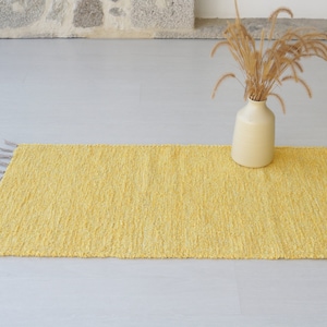 May include: A yellow woven rug with fringe on the edges. The rug is on a light grey floor and a vase with dried flowers is in the background.