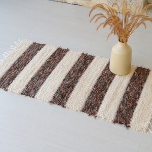 May include: A striped rug with cream and brown bands. The rug has a fringed edge and is made of a textured material. A beige vase with dried plants is nearby. The rug is suitable for a hallway or entryway. The rug measures approximately 120cm in length.