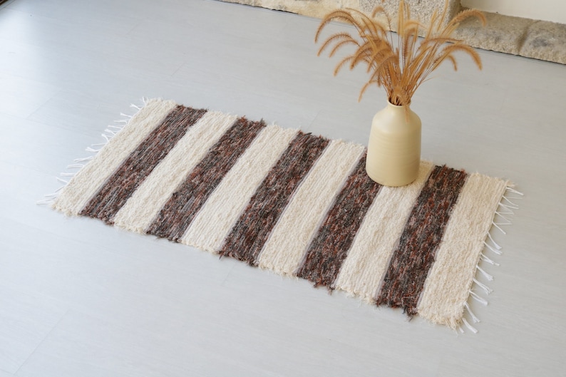 May include: A striped rug with cream and brown bands. The rug has a fringed edge and is made of a textured material. A beige vase with dried plants is nearby. The rug is suitable for a hallway or entryway. The rug measures approximately 120cm in length.