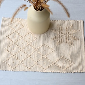 Könnte beinhalten: Ein cremefarbener gewebter Teppich mit einem Rautenmuster und einem Sternendesign. Der Teppich ist aus Naturfasern gefertigt und hat eine strukturierte Oberfläche.
