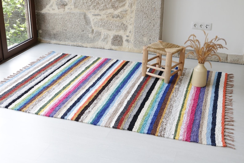 Puede incluir: Una alfombra de rayas coloridas con flecos en los bordes. La alfombra est&aacute; hecha de un material suave y texturizado y tiene una variedad de colores, incluyendo rojo, naranja, amarillo, verde, azul, morado y marr&oacute;n.