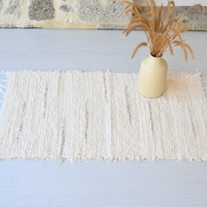 May include: A white, fluffy rug with a textured, woven design. The rug has a frayed edge and is laid out on a light gray floor.