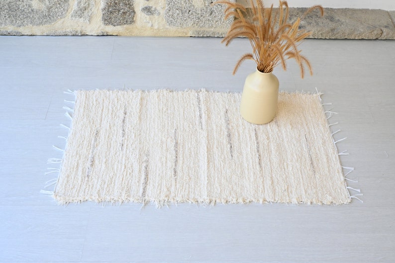 May include: A white, fluffy rug with a textured, woven design. The rug has a frayed edge and is laid out on a light gray floor.