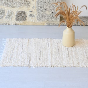 May include: A white woven rug with a fringed edge, placed on a light gray floor. A beige ceramic vase with dried grass stalks sits on the rug.
