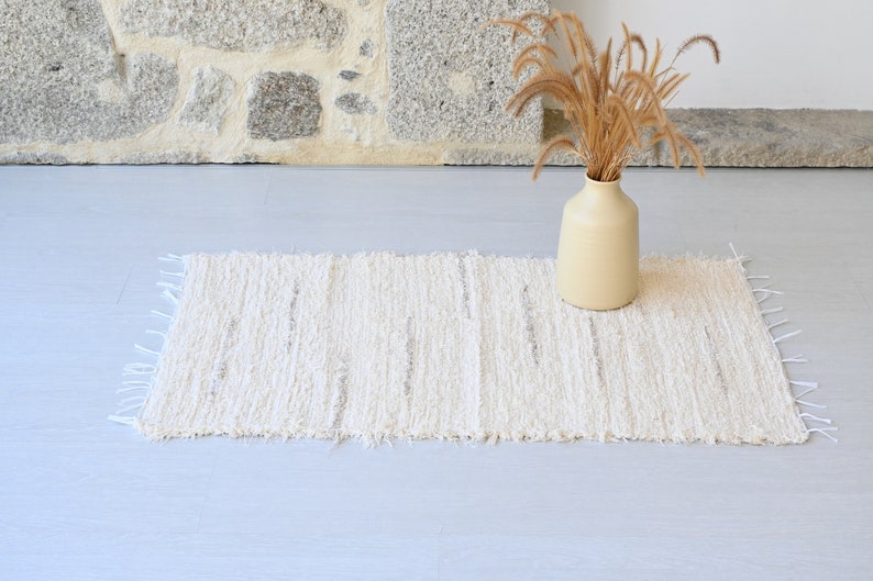 May include: A white woven rug with a fringed edge, placed on a light gray floor. A beige ceramic vase with dried grass stalks sits on the rug.