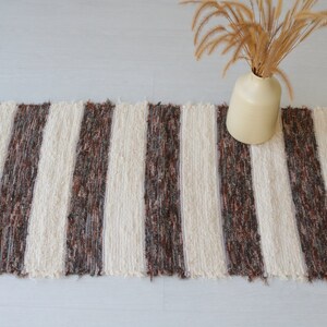May include: A rectangular rug with vertical cream and brown stripes. The rug has a fringed edge and is placed on a light-coloured surface. A beige vase with dried plants sits on the rug.
