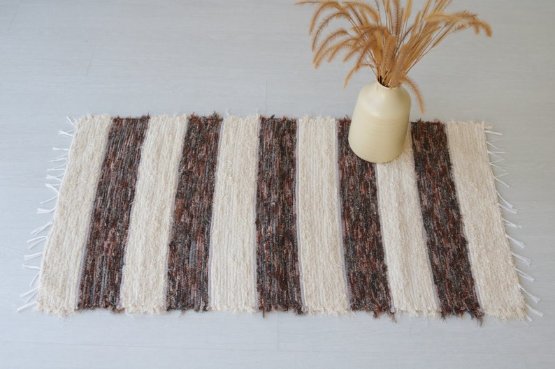 May include: A rectangular rug with vertical cream and brown stripes. The rug has a fringed edge and is placed on a light-coloured surface. A beige vase with dried plants sits on the rug.