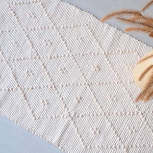 Puede incluir: Camino de mesa de macramé color crema con un patrón de diamantes. El camino de mesa está hecho de hilo grueso y texturizado y presenta pequeños nudos decorativos dentro de cada diamante. Un jarrón con hierba ornamental seca está en la esquina superior derecha.