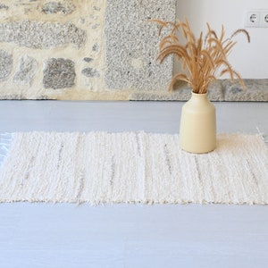 May include: A white woven rug with a fringed edge sits on a light gray floor. A vase with dried grasses sits on the rug.