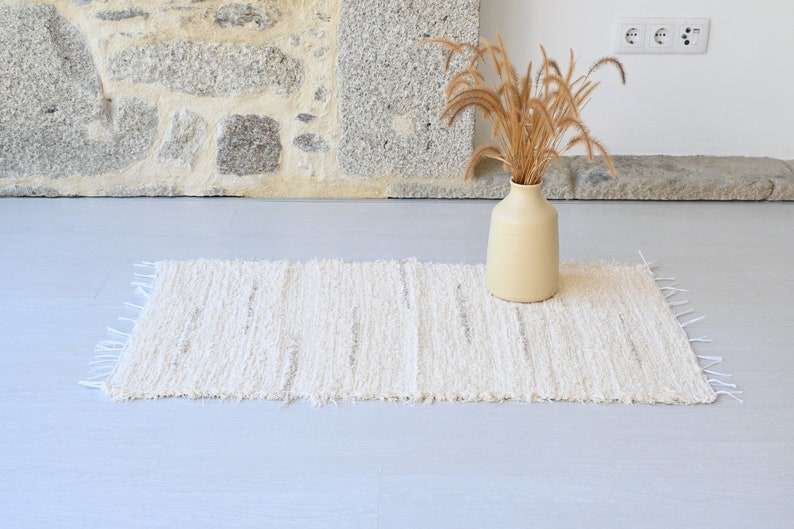 May include: A white woven rug with a fringed edge sits on a light gray floor. A vase with dried grasses sits on the rug.