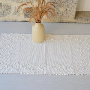 Può includere: Un tappeto rettangolare color bianco sporco caratterizzato da un motivo a rombi testurizzato creato con punti trapuntati in rilievo. Il tappeto ha una superficie a coste ed è mostrato con un vaso in ceramica beige chiaro contenente steli di grano.