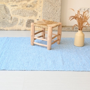 Pu&ograve; includere: Un tappeto shaggy blu chiaro con frange sui bordi. Il tappeto &egrave; su un pavimento bianco e uno sgabello di legno con una seduta intrecciata &egrave; al centro del tappeto.