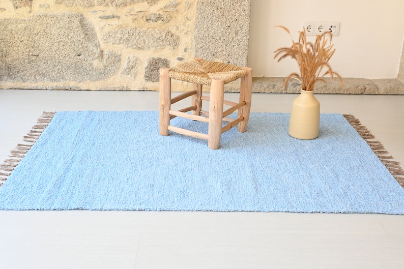 Pu&ograve; includere: Un tappeto shaggy blu chiaro con frange sui bordi. Il tappeto &egrave; su un pavimento bianco e uno sgabello di legno con una seduta intrecciata &egrave; al centro del tappeto.