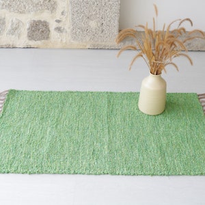 May include: A green woven rug with fringe on the edges. The rug is on a white floor and there is a vase with dried grass in the background.