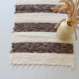 May include: A woven rug with alternating cream and brown horizontal stripes. The rug has a fringed edge and is paired with a beige vase holding dried ornamental grass. The rug is a decorative home accent.