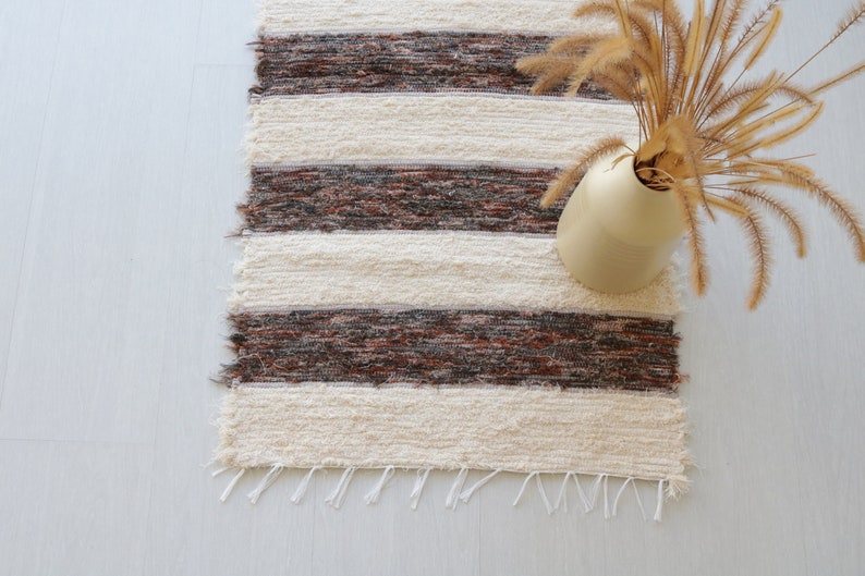 May include: A woven rug with alternating cream and brown horizontal stripes. The rug has a fringed edge and is paired with a beige vase holding dried ornamental grass. The rug is a decorative home accent.