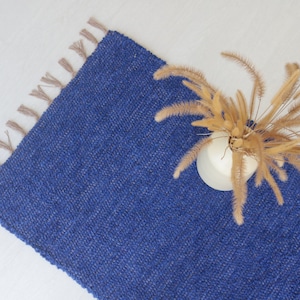 May include: A blue woven rug with fringe on the edges. The rug is lying flat on a white surface with a vase of dried flowers on top of it.