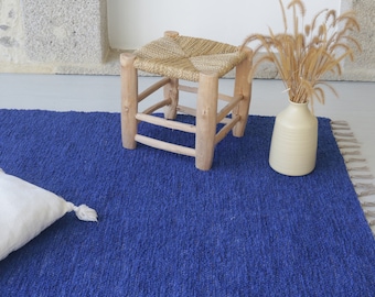 Puede incluir: Una vibrante alfombra azul con tejido texturizado, un pequeño taburete de madera con asiento tejido y un jarrón crema con hierbas secas. Una almohada blanca con borlas descansa sobre la alfombra. La escena evoca una estética acogedora y natural.