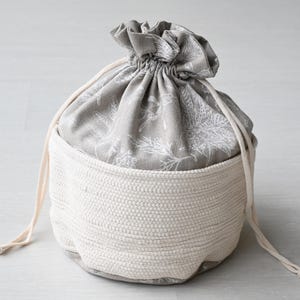 Può includere: Una borsa portaoggetti rotonda bicolore. Il fondo è in materiale bianco sporco intrecciato, mentre la parte superiore è in tessuto grigio con un motivo floreale bianco. Ha una chiusura con coulisse con cordoncini bianchi sporchi abbinati.