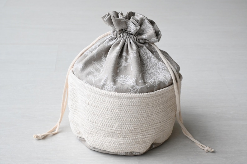 Pu&ograve; includere: Una borsa portaoggetti rotonda bicolore. Il fondo &egrave; in materiale bianco sporco intrecciato, mentre la parte superiore &egrave; in tessuto grigio con un motivo floreale bianco. Ha una chiusura con coulisse con cordoncini bianchi sporchi abbinati.