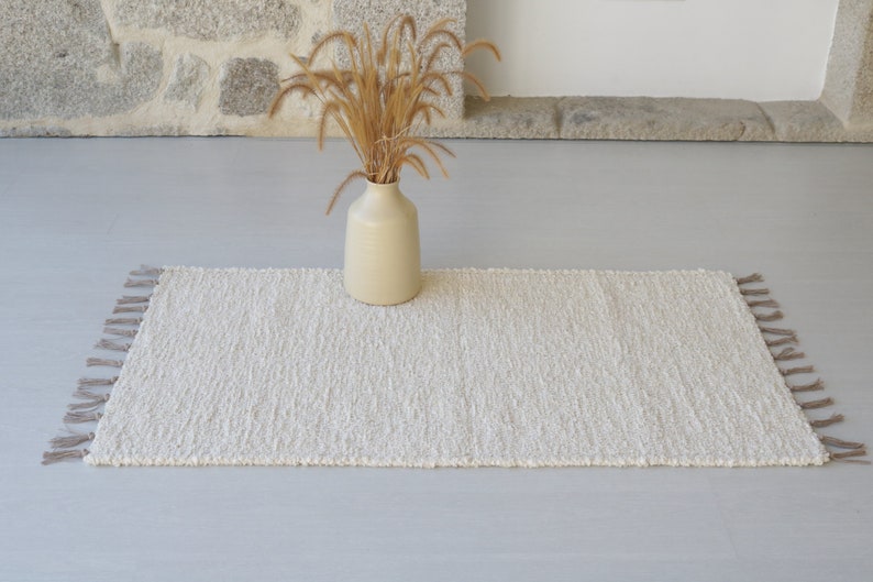 Puede incluir: Una alfombra blanca y esponjosa con flecos en los bordes. La alfombra est&aacute; colocada sobre un suelo gris claro con un jarr&oacute;n de hierba seca en el centro.