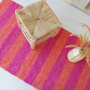 May include: A pink and orange striped rug with a woven stool and a vase of dried grass.