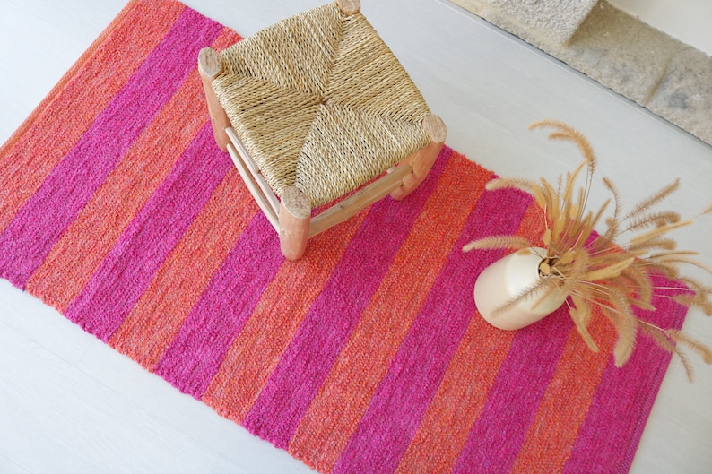 May include: A pink and orange striped rug with a woven stool and a vase of dried grass.