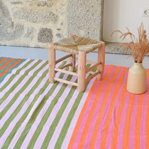 Op de afbeelding: Een rechthoekig geweven textiel met verticale strepen in afwisselende kleuren oranje, lichtroze en olijfgroen. De stof heeft een gefranjerde rand en wordt getoond naast een kleine houten kruk met een gevlochten zitting en een beige keramische vaas.