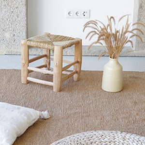 Könnte beinhalten: Ein Naturfaser-Teppich mit gewebter Struktur in hellbrauner Farbe. Ein Holzhocker mit gewebtem Sitz befindet sich auf dem Teppich. Eine Vase mit getrocknetem Gras steht auf dem Teppich.