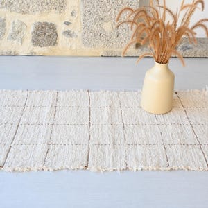 Puede incluir: Una alfombra rectangular tejida en color crema con un patrón de cuadrícula y bordes con flecos. Un jarrón beige contiene hierba ornamental seca. La alfombra está sobre una superficie gris claro, con una pared de piedra al fondo.