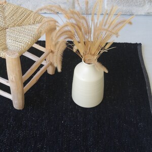 May include: A black woven rug with a white border. A wooden stool with a woven straw seat sits on the rug. A vase with dried grass sits on the rug.