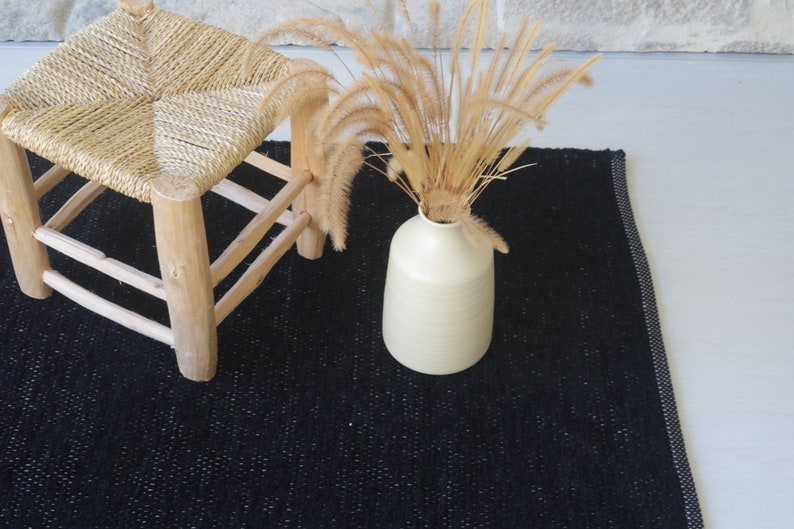 May include: A black woven rug with a white border. A wooden stool with a woven straw seat sits on the rug. A vase with dried grass sits on the rug.
