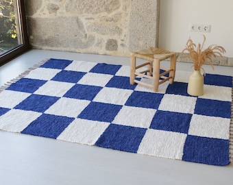 Könnte beinhalten: Ein blau-weiß karierter Teppich mit Fransen. Der Teppich besteht aus einem gewebten Material und hat ein sich wiederholendes Muster aus großen Quadraten. Ein kleiner Holzschemel und eine Vase mit getrockneten Pflanzen befinden sich auf dem Teppich.