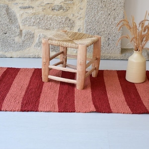 Op de afbeelding: Een rechthoekig kleed met afwisselende verticale strepen in tinten dieprood, baksteenrood en gedempt koraal. Het kleed ligt op een lichte ondergrond, met een kleine houten kruk met een gevlochten zitting en een beige keramieken vaas ernaast.